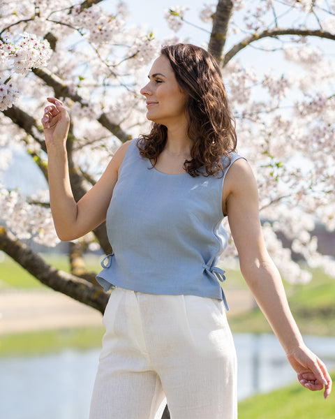 A woman with long, wavy brown hair wears a cloudy blue sleeveless top with side ties and white linen pants, looking off to the right. She stands outdoors under a blooming cherry blossom tree, with a calm body of water and green landscape in the background.