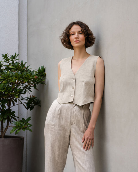 A light-skinned woman with short, curly brown hair stands confidently against a textured light gray wall. She is wearing a matching menique linen outfit in the color natural, consisting of a cropped, sleeveless vest and high-waisted, wide-legged trousers with pleats at the front. Her posture is relaxed yet poised, with one hand slightly tucked into her pocket.