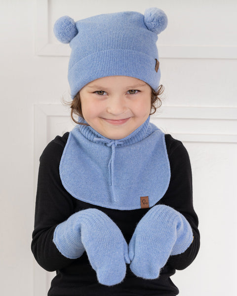 A young child with light brown curly hair and a subtle smile, is looking directly at the viewer. They are wearing a light blue knit hat with two light blue pom-poms on top, a matching light blue knit neck warmer with a tie, and light blue mittens, over a black long-sleeved shirt. The background is a white paneled wall.
