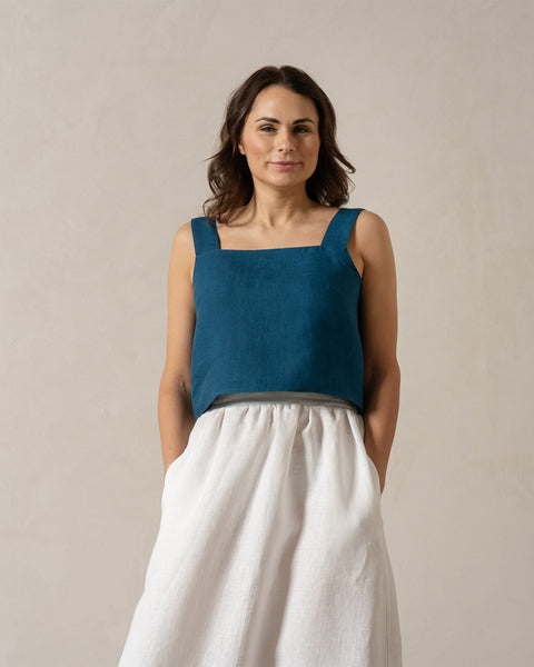 A woman with medium-toned skin and dark hair looks directly at the camera with a neutral expression. She is wearing a teal, sleeveless square-neck linen top and a pure white, menique
linen skirt, with her hands in the skirt's pockets. The background is a plain, light beige color.
