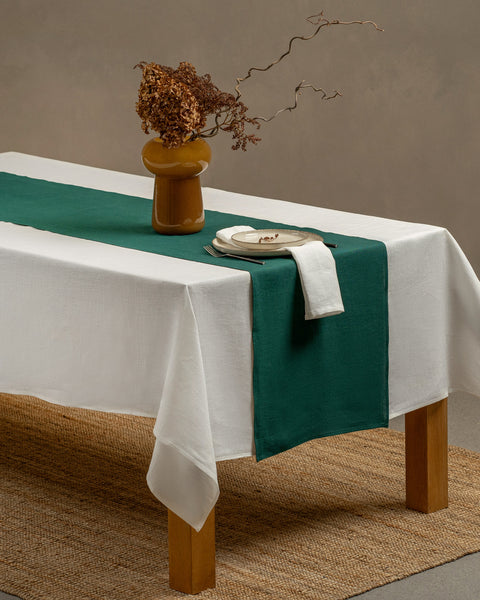 The menique linen tablecloth in pure white color and table runner in dark green color, draping over a wooden table, with a glass vase and a teacup with a plate on top of it.