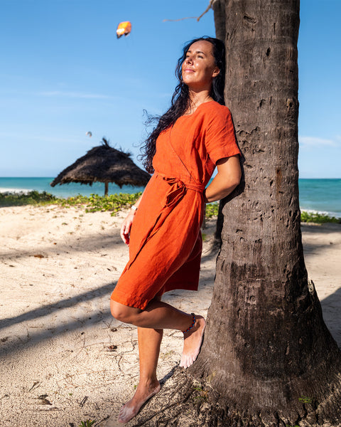 A smiling woman with long dark hair stands barefoot on a sandy beach, holding a wide-brimmed hat above her head with both hands. She is wearing a cinnamon red linen sundress with thin straps. The ocean is in the background with gentle waves, and several boats are visible in the distance under a bright sky.