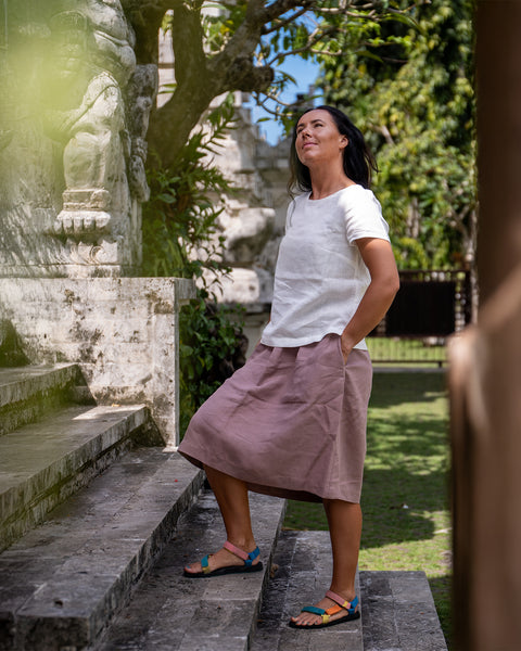 A woman with dark hair stands on stone steps outdoors, looking upwards. She is wearing a white short-sleeved top, a dusty pink knee-length skirt, and colourful flat sandals. Her hands are in her skirt pockets. Beside her is a weathered stone wall with carvings, and trees with green leaves are visible in the background under a bright sky.