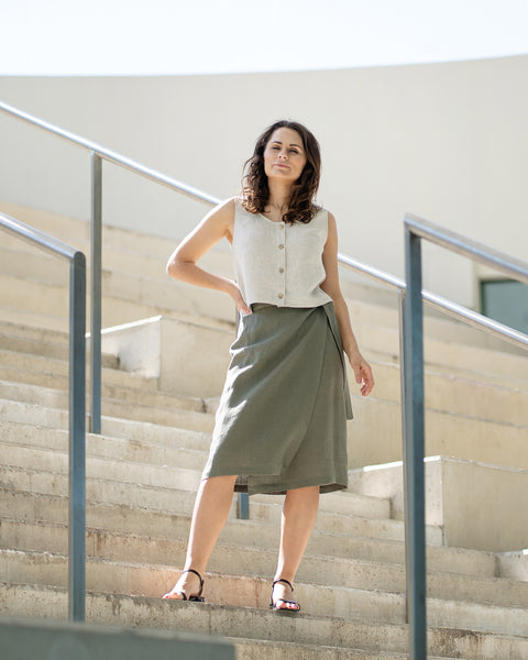 A woman with brown hair, wearing a natural linen button-front sleeveless top and a stone green wrap skirt, poses on outdoor concrete stairs.