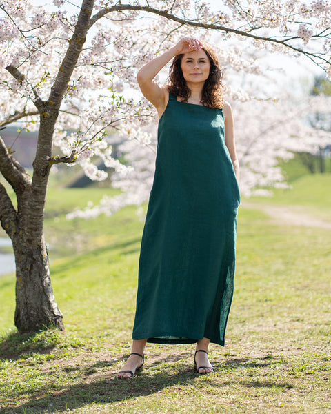 A woman with brown hair, wearing a long, dark green, sleeveless maxi dress, stands outdoors next to a tree with white blossoms, shading her eyes.
