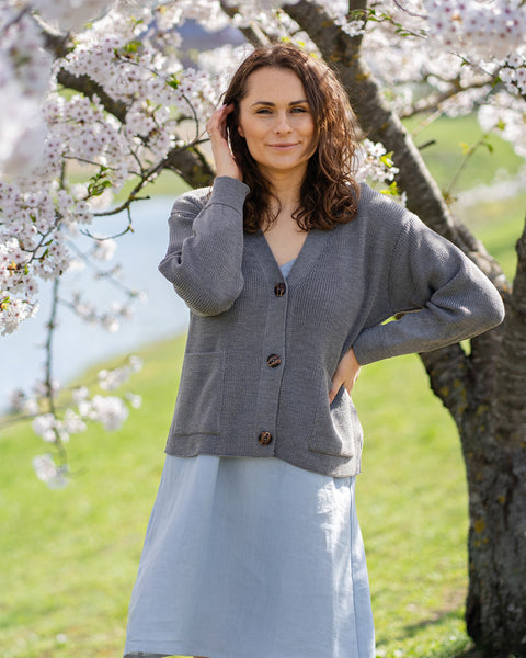 A woman with brown hair, wearing a grey knit cardigan over a light blue dress, stands outdoors next to a tree with white blossoms.