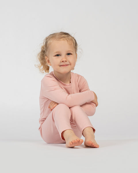 Little girl sitting barefoot in a pink bamboo kids’ clothing set with long sleeves and pants, smiling against a white background.