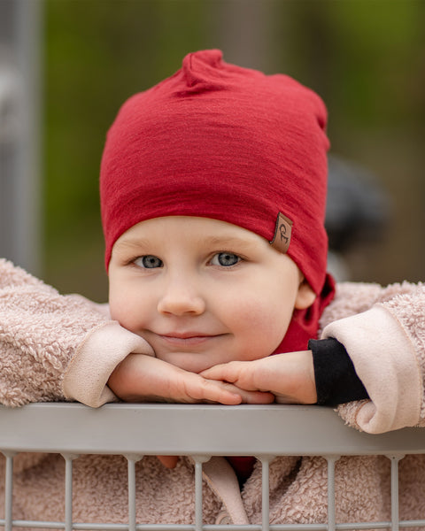 Close-up of child resting on playground railing, wearing a red Merino wool beanie and neck gaiter, cozy outdoor setting.