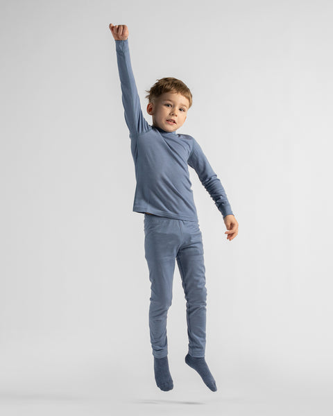 Full-length view of a young boy posing playfully with one arm raised in the air, dressed in matching dusty blue base layer top and leggings.