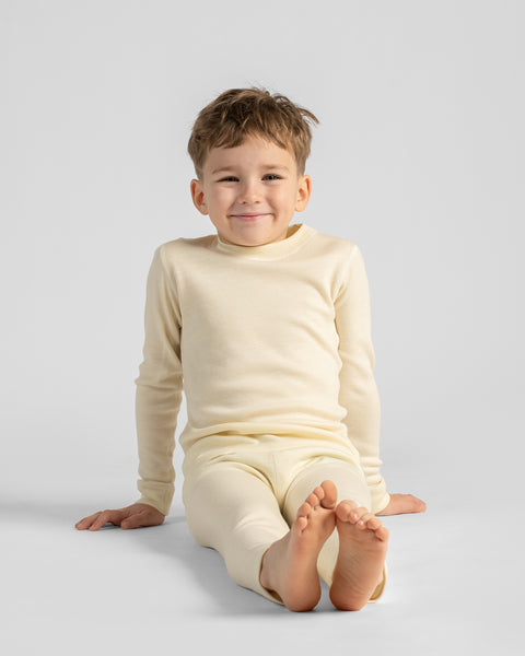 Young boy sitting comfortably barefoot on the floor, wearing an off-white long-sleeved thermal base layer shirt and pants.