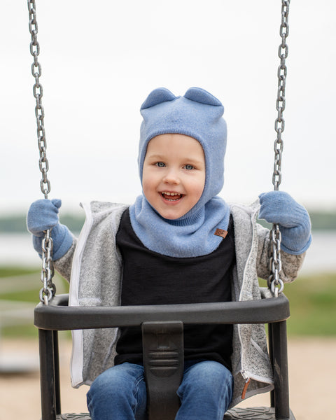 A young child with light skin and light hair is sitting on a swing at a playground. The child is wearing a light blue balaclava-style hat with ear-like protrusions, matching light blue mittens, a dark long-sleeved shirt, and a gray zippered hoodie.