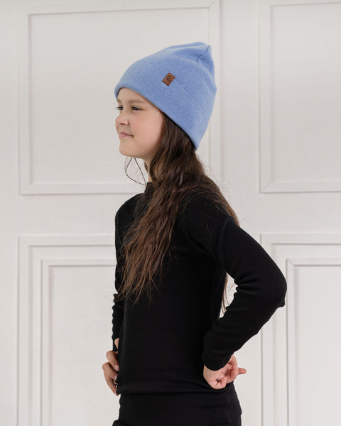 A child with long dark brown hair, is seen in profile, looking to the left. They are wearing a light blue knit beanie with a small brown label and a black long-sleeved shirt. Their hands are behind their back, and the background is a white paneled wall.