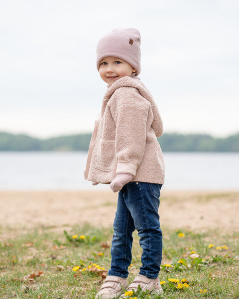A young child with a slight smile looking over their shoulder at the viewer. They are standing on grassy ground with dandelions, wearing a light pink knit beanie, a beige sherpa coat, blue jeans, and pink sneakers, with a blurred body of water and sky in the background.