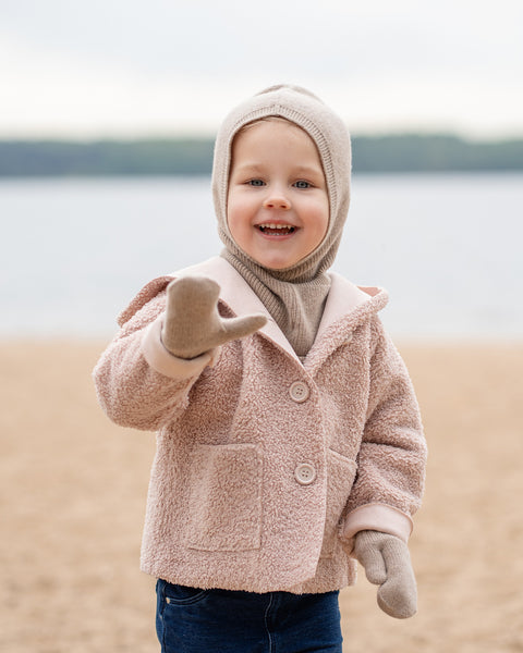 A cheerful young child with blue eyes and an open-mouthed smile, stands on a sandy beach, extending one hand towards the viewer. They are wearing a beige balaclava, a matching beige scarf, a light pink sherpa-style coat, beige mittens, and blue jeans, with a blurred body of water and sky in the background.