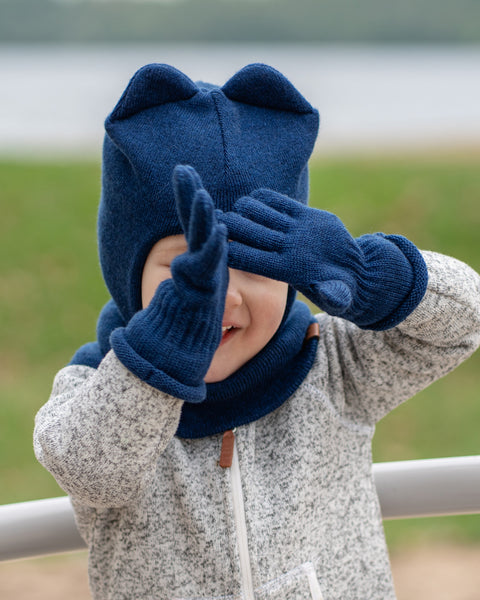 A young child, appearing to have light-toned skin, is seen from the chest up, playing outdoors. They are wearing a grey speckled zip-up jacket, a dark blue knitted menique balaclava-style hat with two rounded ears on top, and matching dark blue knitted gloves. The child's hands are raised to cover their eyes, as if playing peek-a-boo, but a wide smile is visible below their hands. A light-colored railing is in the foreground, and a blurred natural background with water is behind them.
