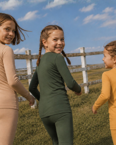 Three children walking away through a grassy field while looking back over their shoulders and smiling, wearing two-piece, long-sleeved thermal sets in beige, dark green, and mustard yellow.