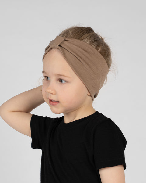 A side profile portrait of a young girl looking off to the left against a plain light grey background. She is wearing a black short-sleeved t-shirt and a beige ribbon headband. One hand rests lightly behind her head.