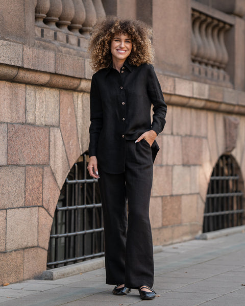 A smiling woman with voluminous, light brown curly hair stands in front of a textured stone building. She is wearing a pure black menique linen shirt tucked into wide-legged black linen pants and black shoes, with one hand casually placed in her pocket.