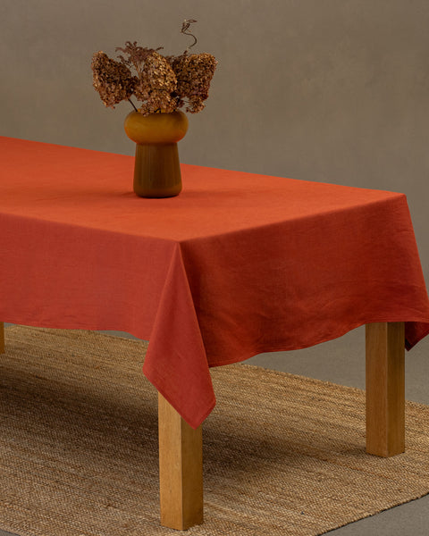 The menique linen tablecloth in cinnamon red color, draping over a wooden table, with a vase on top of it.