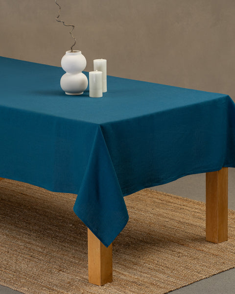 The menique cobalt blue linen tablecloth on a wooden table. On top of the tablecloth is a vase and candles. Under the table is a carpet.