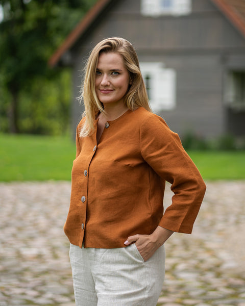 A blonde woman smiles warmly at the camera while standing outside a rustic wooden house. She is wearing a long-sleeved orange shirt and light-colored pants.
