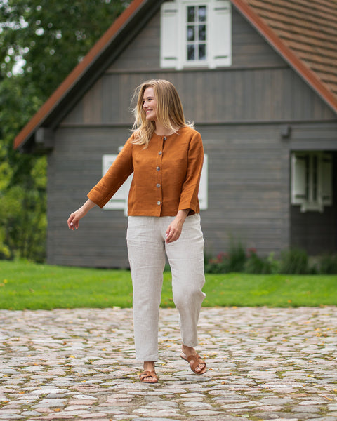 A blonde woman in a rust-colored top and light trousers poses playfully on a gravel driveway with her arms slightly outstretched. Behind her is a traditional dark wooden house surrounded by a green lawn.