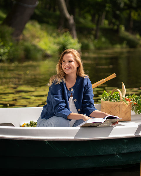 A blonde woman smiles brightly while sitting in a rowboat surrounded by green trees. She is dressed in a blue jacket and light-colored top while enjoying a day on the water.