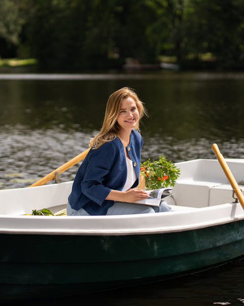 A woman in a blue jacket sits sideways in a small rowboat on a calm body of water. She looks down thoughtfully with wooden oars resting near her in the boat.