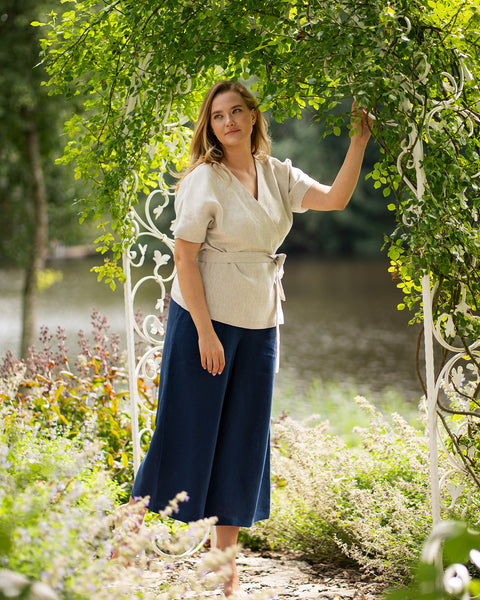 A woman stands under a leafy white garden archway near a river, resting one hand lightly on the structure while gazing off into the distance. Her outfit features a natural beige, short-sleeved linen wrap top tied at the waist and storm blue wide-leg pants.
