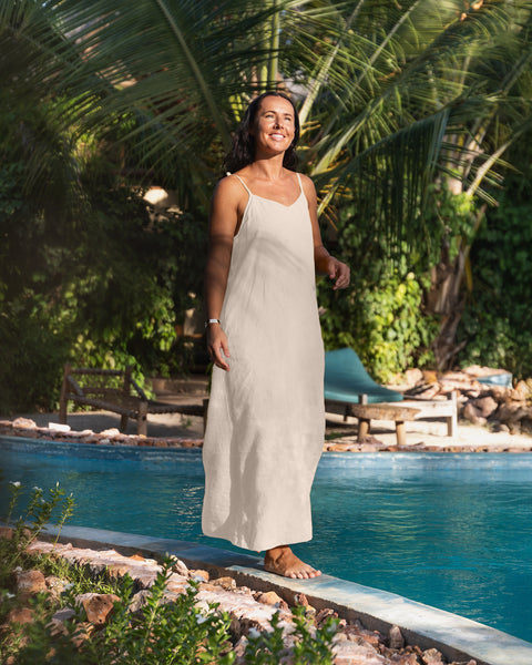 A woman with dark hair wearing a natural linen sundress. Behind her is a bright blue pool surrounded by palm trees and tropical foliage. The ground is light-colored with shadows.