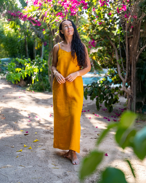 A woman with long, dark, wavy hair stands barefoot on a sandy path, looking up towards the left. She is wearing a mustard yellow linen sundress with thin straps. Lush green plants and trees surround her, and there are pink flowers blooming in the background. Yellow petals are scattered on the path.