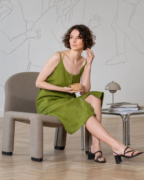 A woman with short, curly brown hair, wearing a sleeveless, button-front, forest green midi-dress and black block-heeled sandals, sits in a modern gray armchair. She holds a small, light brown cup in her right hand and looks off to the left with a pensive expression. Her left leg is crossed over her right, and her left hand is touching her chin.