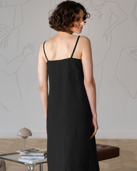 A shot from behind and slightly to the right shows a woman with short, curly dark hair, wearing a sleeveless, square-neck, black dress with thin straps. She is looking to her right. A light-colored wall with faint white line art is in the background, and a small, gray dome-shaped lamp on a glass table is partially visible in the lower left corner.