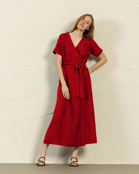 A person with long reddish-blonde hair poses against a textured off-white wall. They are wearing a long-sleeved red wrap dress with a tie at the waist and black sandals.