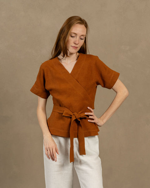 A fair-skinned person with long, auburn hair, wearing V-neck linen wrap top with short, wide sleeves and light-colored pants. They are holding the ends of the top's tie in front of them, forming a bow.