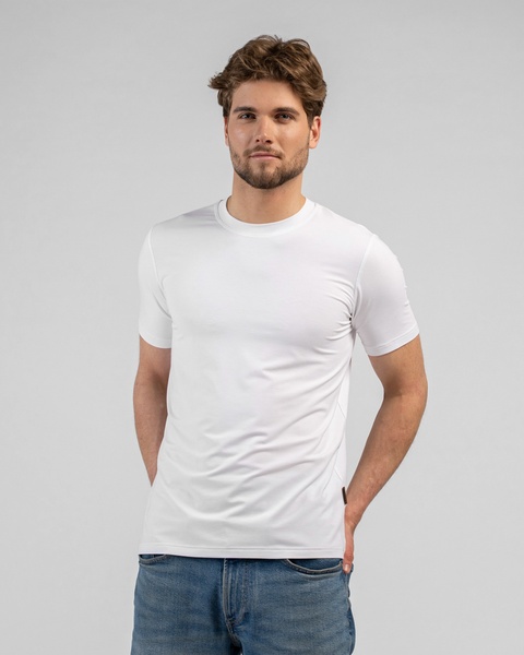 A studio portrait of a man looking directly at the camera with his hands resting behind his back. He is wearing a classic white short-sleeved t-shirt and blue jeans against a light grey background.