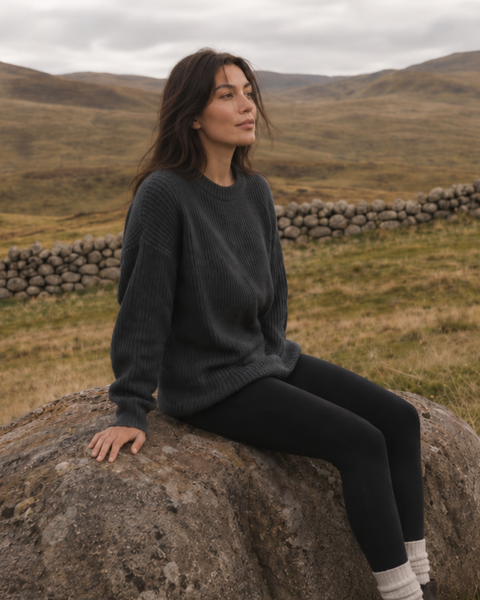 A woman sits calmly on a large boulder in a sprawling, grassy landscape with rolling hills. She is wearing an oversized dark grey ribbed sweater, black leggings, and light-colored socks, looking off toward a low stone wall in the distance under a cloudy sky.