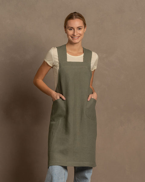 A woman with her hands in the pockets of a menique stone green linen cross-back apron, wearing a light blue jeans, stands smiling against a brown textured background.