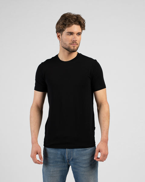 A studio portrait of a man looking off to the right side of the frame with his arms resting by his sides. He is wearing a solid black short-sleeved t-shirt and medium-wash blue jeans.