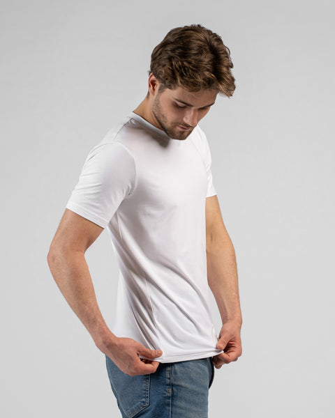 A man stands looking downwards while holding the bottom hem of his white short-sleeved t-shirt with both hands. He is wearing blue jeans against a plain light grey background.