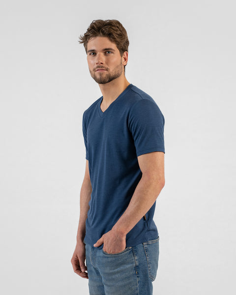 A side-profile studio portrait of a man looking directly at the camera. He is wearing a denim blue v-neck t-shirt and blue jeans with his left hand in his pocket.