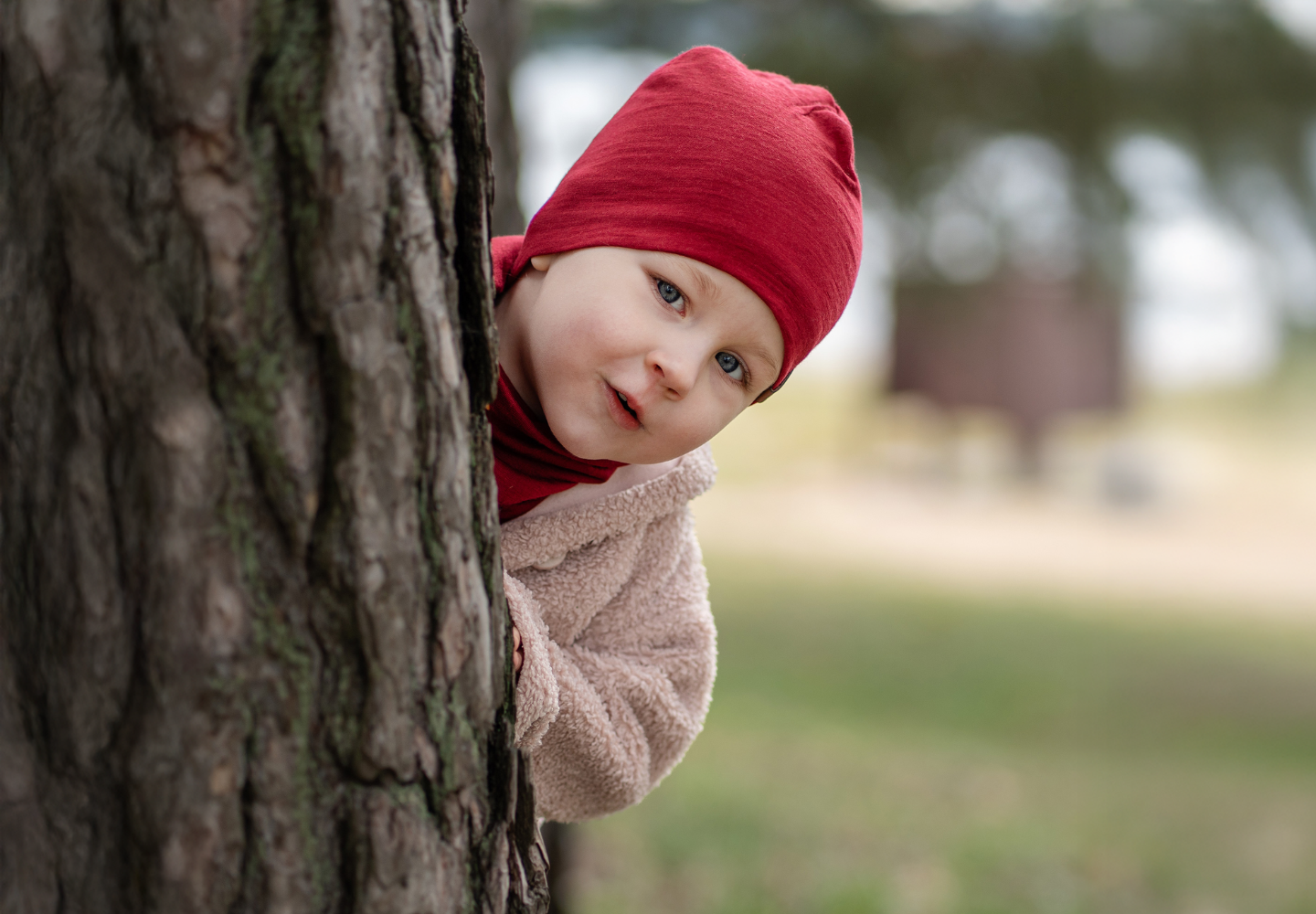 Kid wearing red merino wool beanie and neck gaiter peeking from behind a tree outdoors in autumn nature.