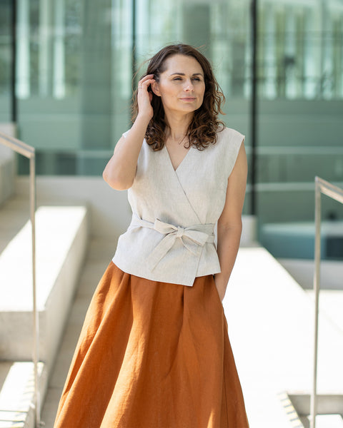 A woman with shoulder-length, wavy brown hair poses outdoors, looking slightly to her right. She wears a natural linen V-neck wrap top with a tie closure at the waist, paired with a long menique linen skirt.