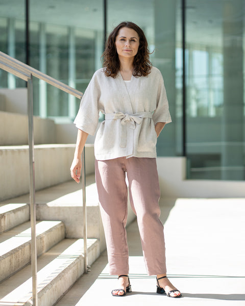 A woman with brown hair, wearing a light beige kimono-style jacket over mauve trousers, stands on indoor concrete stairs.