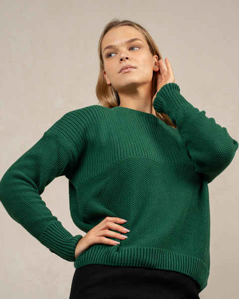 A woman captured from below at an angle, wearing the menique combed cotton sweater in green color. She has one of her hands on her hip, and the other is brushing lightly against the side of her face. The sweater is knitted in a honeycomb stitched.