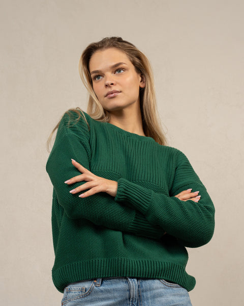 A woman captured from an angle from below, wearing the menique combed cotton sweater SONATA in green color. She has her arms crossed over her waist.