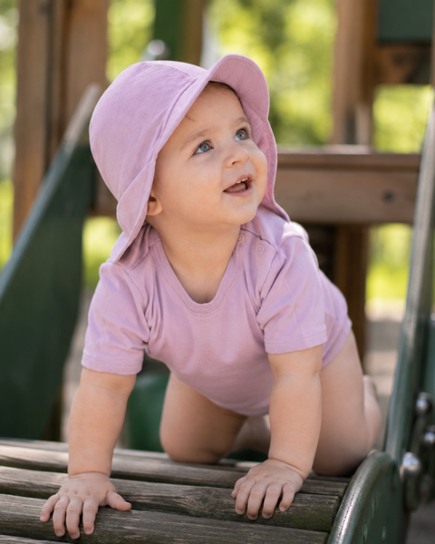 A smiling baby crawling on a wooden playground structure outdoors, wearing a light pink short-sleeve bodysuit and a matching dusty pink sun cap with a wide brim and neck flap.