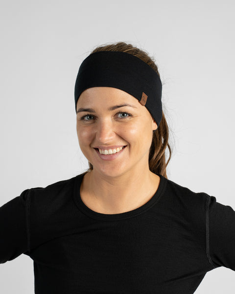 A chest-up studio portrait of a woman smiling forward with hands on her hips, wearing a black top and a black knit headband.
