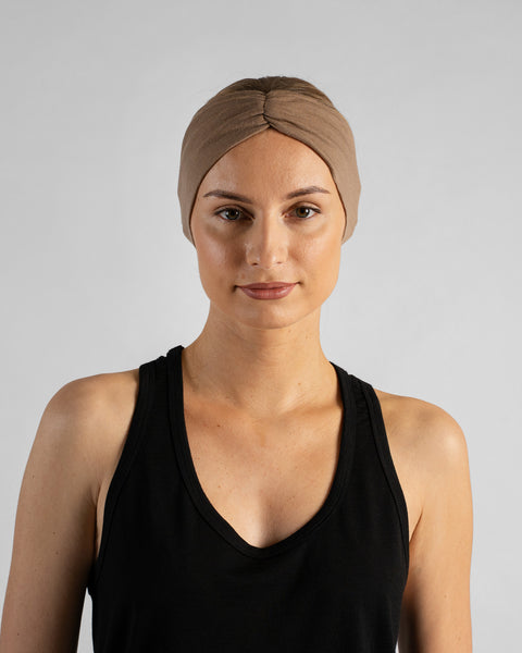 A studio portrait of a blonde woman looking directly at the camera with a soft smile against a plain light grey background. She is wearing a black sleeveless top and a beige merino ribbon headband with the twisted ribbon detail centered on her forehead.