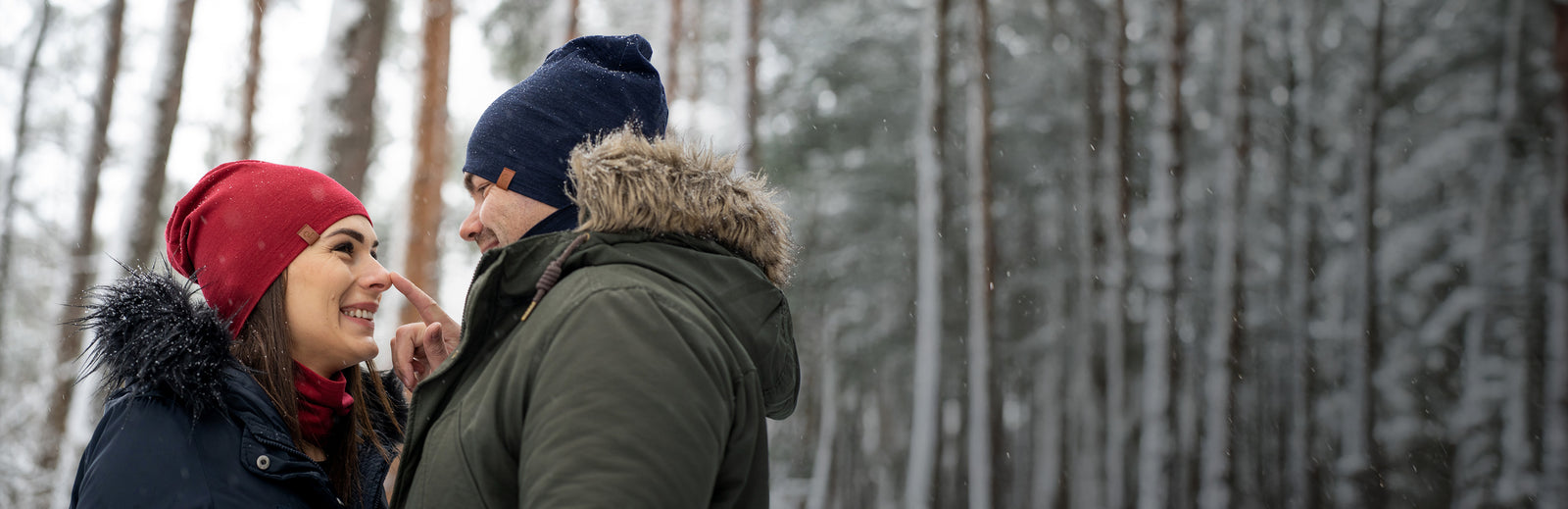 Smiling couple wearing winter hats and jackets, standing close together in a snowy forest